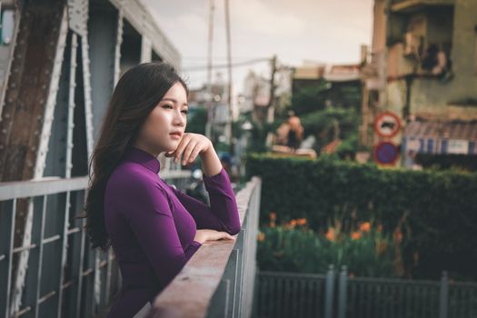 白天，一位身穿紫色连衣裙的亚洲女子在城市桥梁上若有所思地摆着姿势