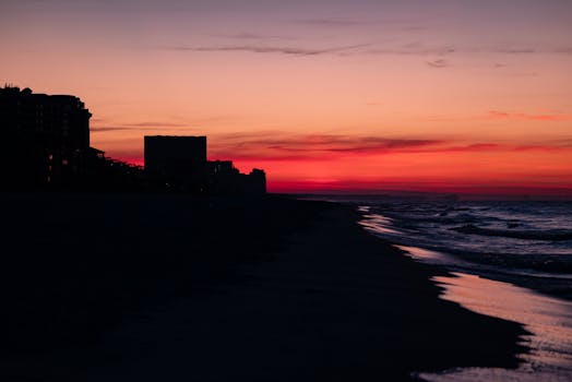 Stunning silhouette of coastal buildings against a vibrant sunset sky, with waves gently lapping the shore.