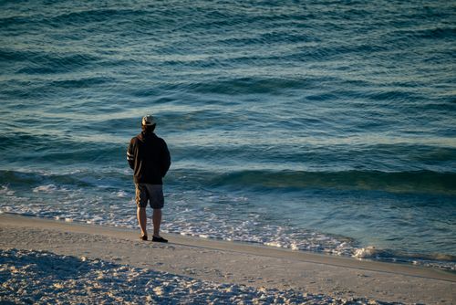 Free A solitary man stands on a tranquil beach, gazing at the sea, evoking a sense of calm and reflection. Stock Photo