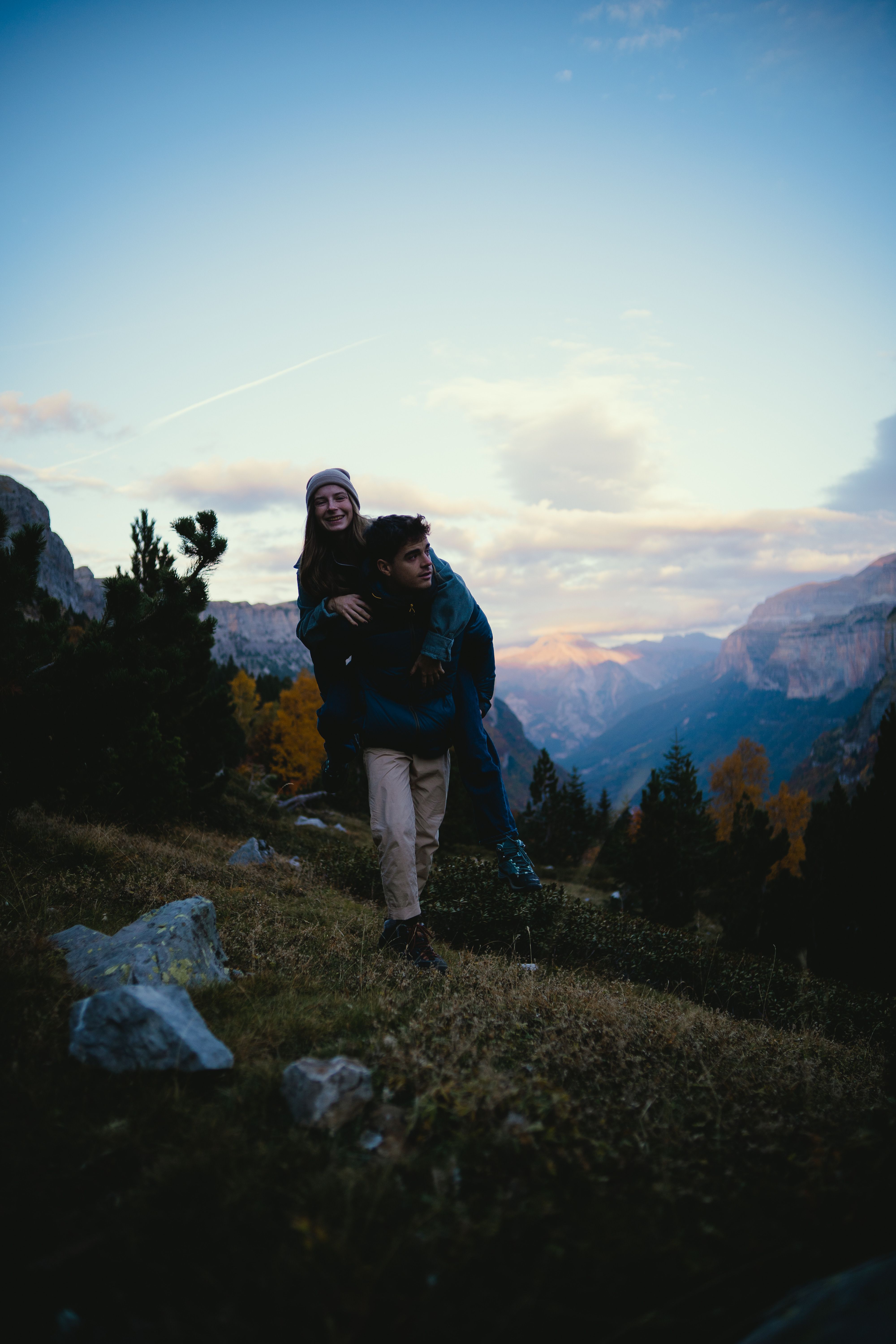 A couple enjoys a piggyback ride on a mountain trail, surrounded by stunning natural scenery.
