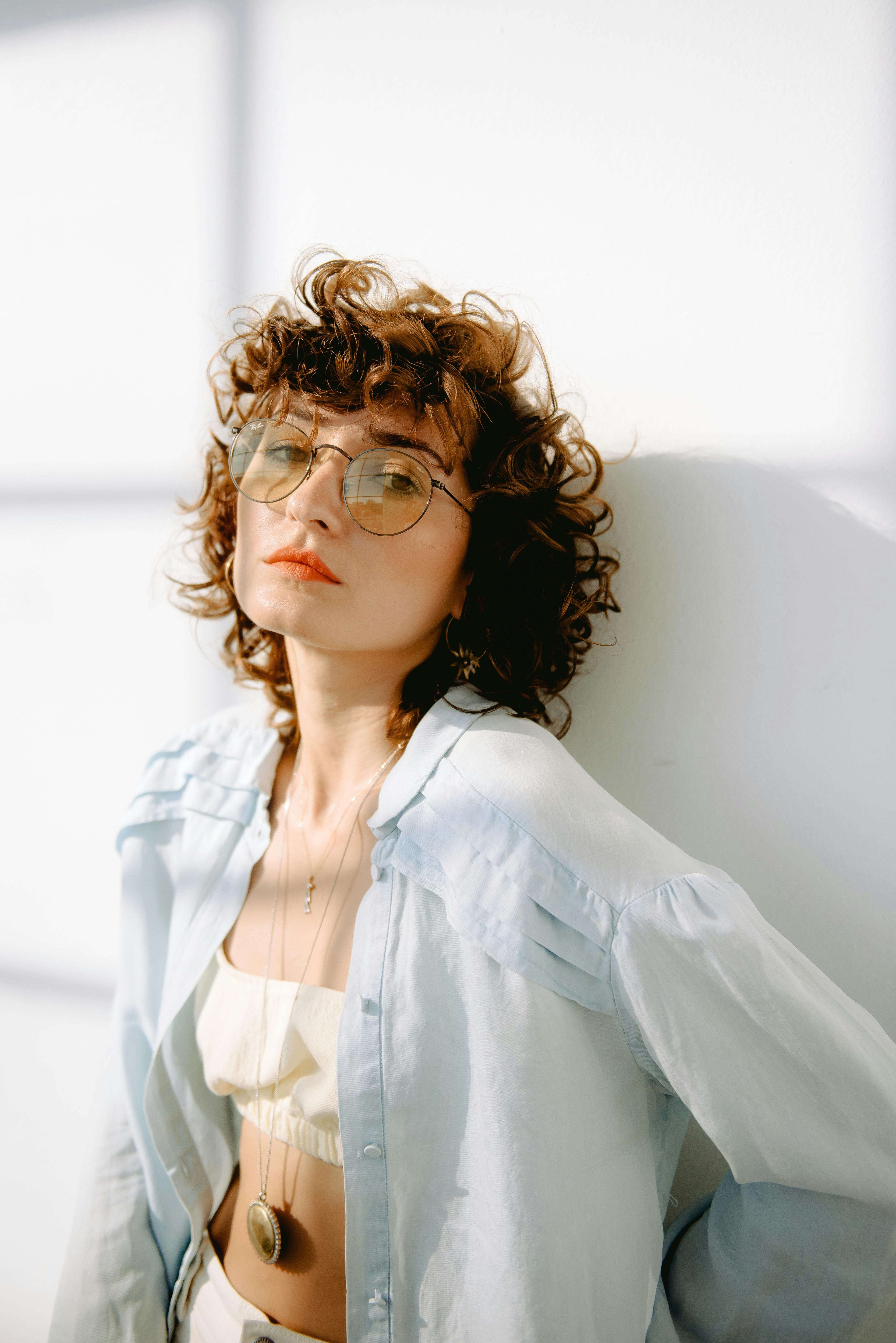Portrait of a young woman with curly hair and sunglasses, posing against a white background.