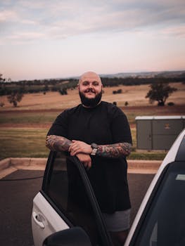 A bald man with tattoos smiling while standing by an open car door in a rural setting.