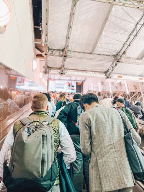 A bustling crowd moves up an escalator in a busy indoor environment, with a focus on diverse attire and backpacks.