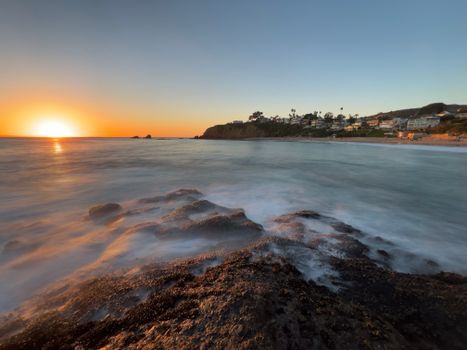 Stunning sunset over a rocky coastline with gentle ocean waves and distant shoreline view.