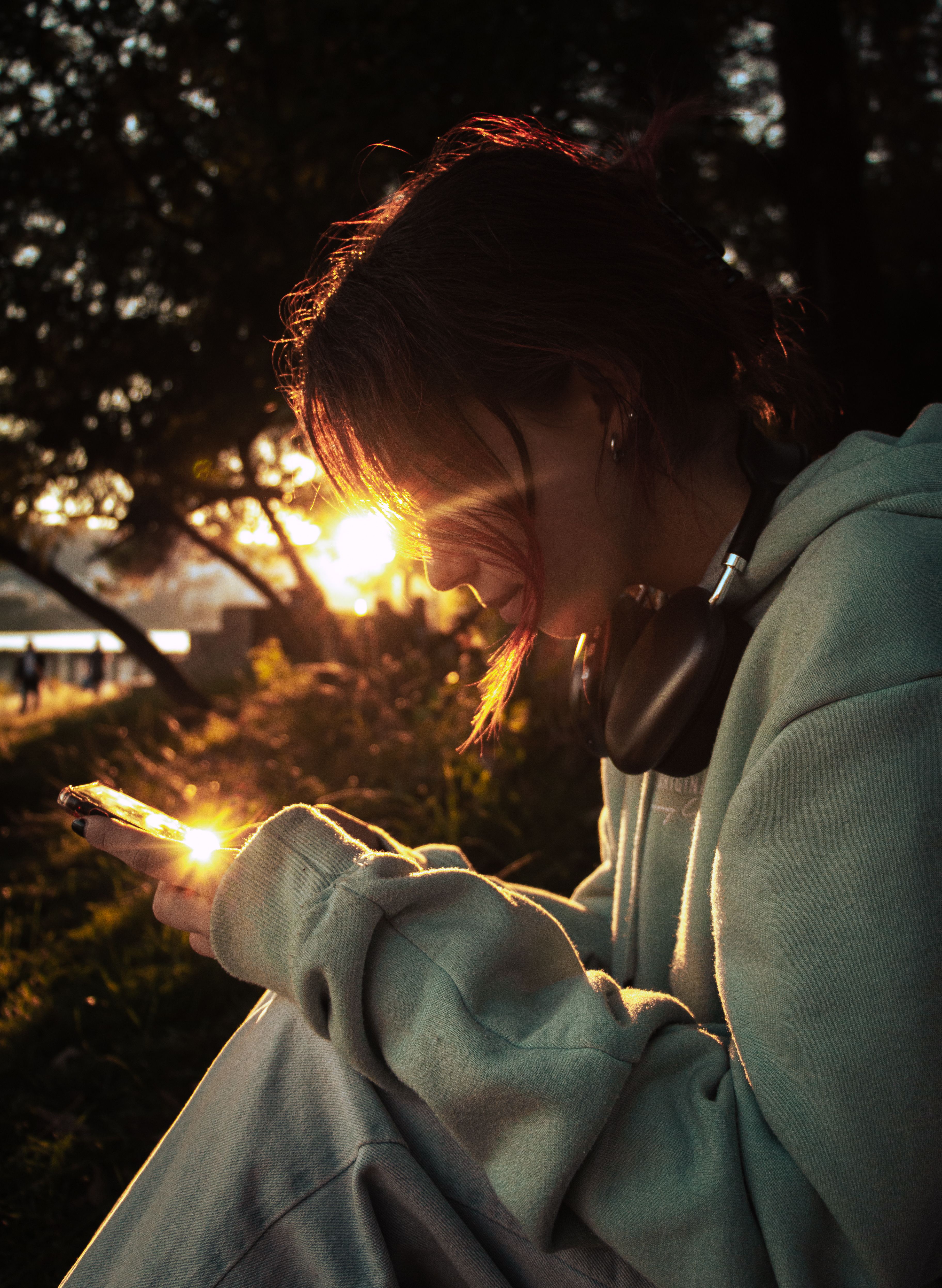 A woman wearing headphones and a hoodie relaxes outdoors with her smartphone at sunset in Tirana, Albania.