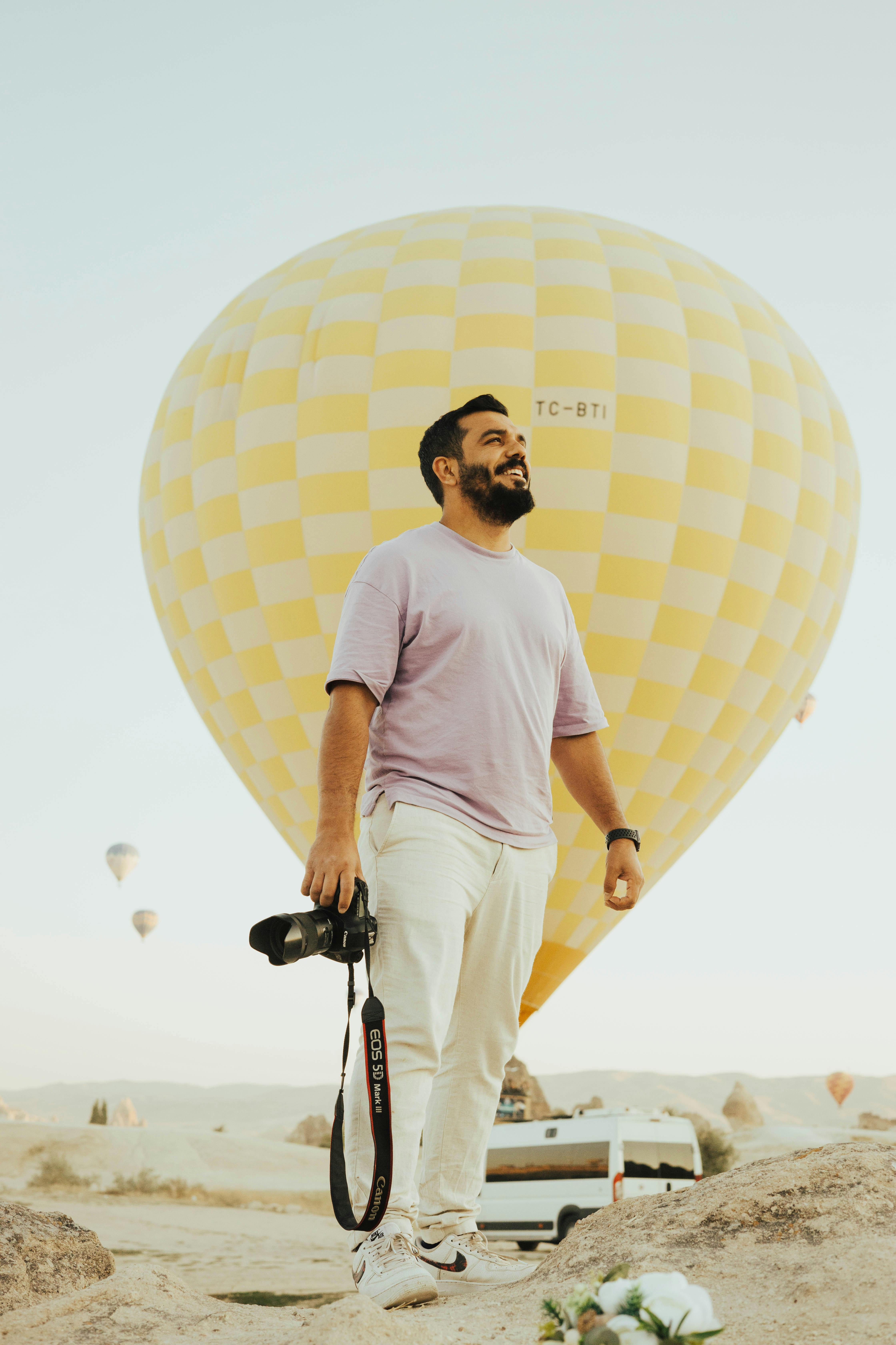 A smiling man with a camera stands by a hot air balloon in a scenic outdoors setting.