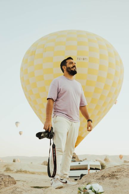 A smiling man with a camera stands by a hot air balloon in a scenic outdoors setting.