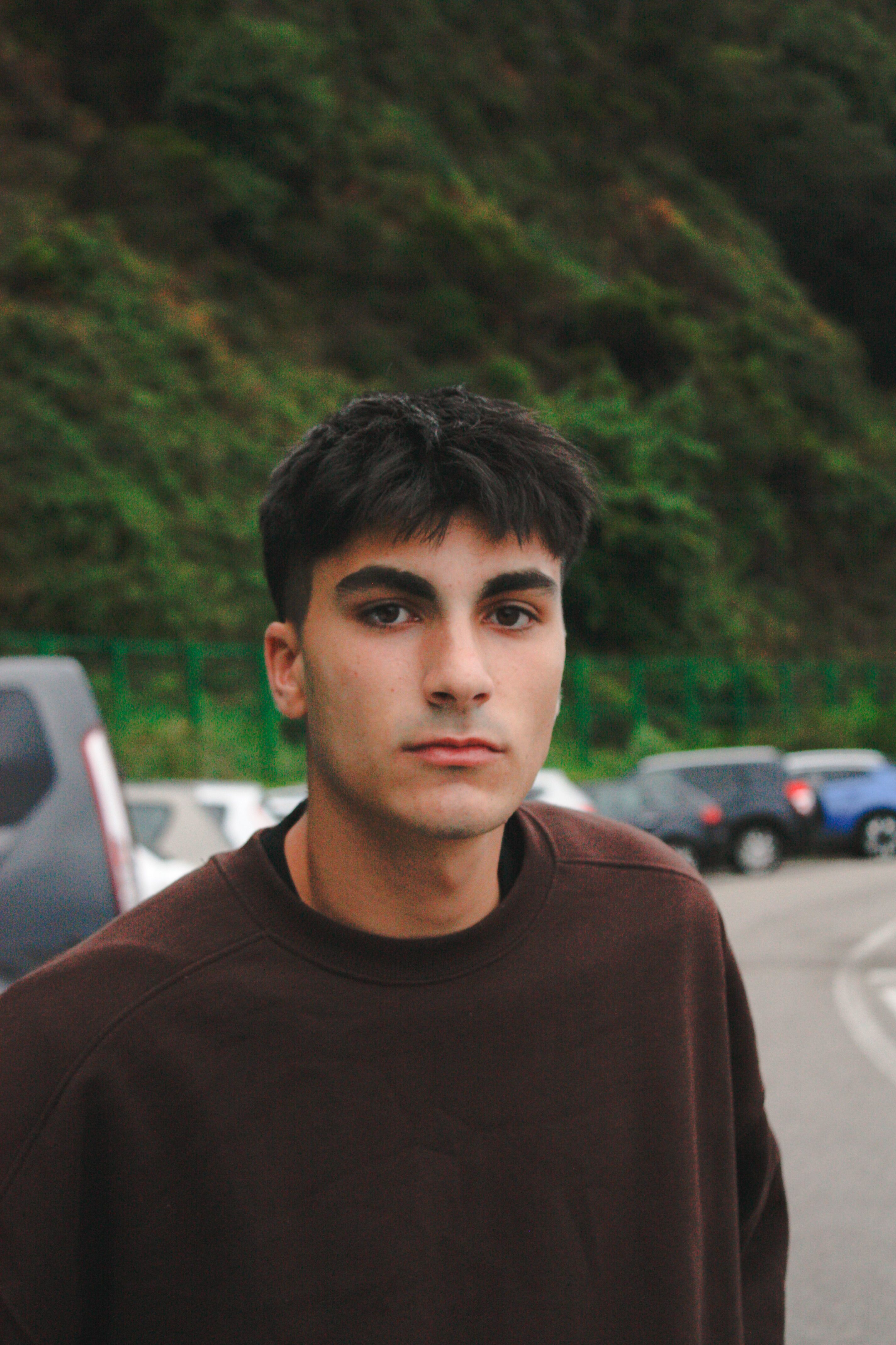 Portrait of a young man in a brown sweater outdoors with parked cars in background.