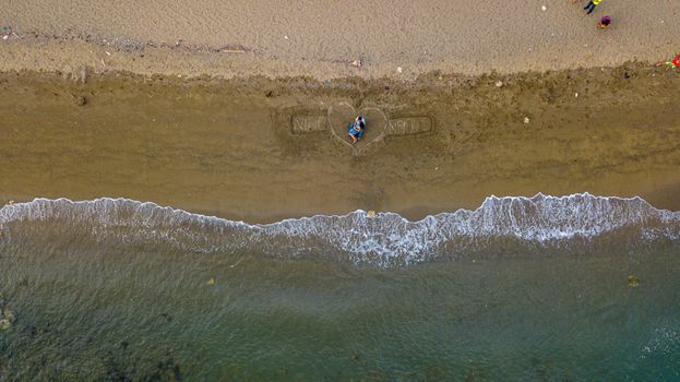Romantic aerial shot of a couple on a beach with heart drawing in sand near the shoreline.