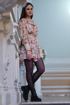 A young woman leans elegantly in a floral dress, indoor setting.