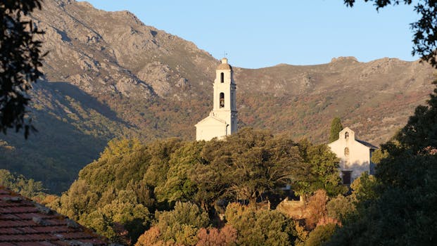 Charming church nestled in lush Corsican landscape, set against scenic mountains under a clear sky.