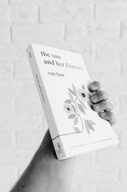 Black and white photo of a hand holding 'The Sun and Her Flowers' by Rupi Kaur.