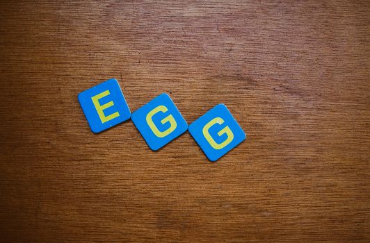 Blue and yellow letter tiles spelling 'EGG' on a wooden surface, in a still life composition.
