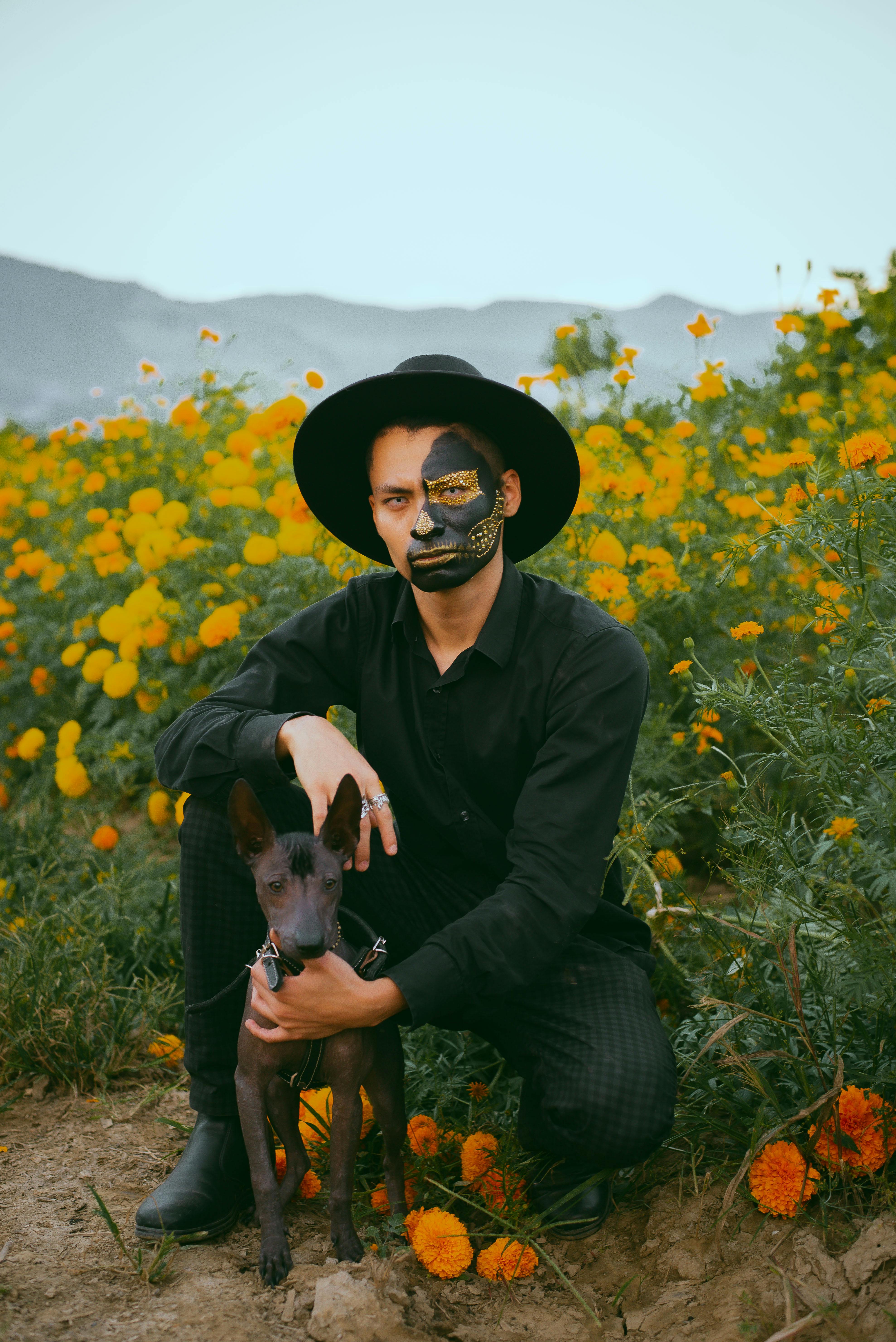 A man with artistic face paint and a dog in a vibrant marigold field during the day.