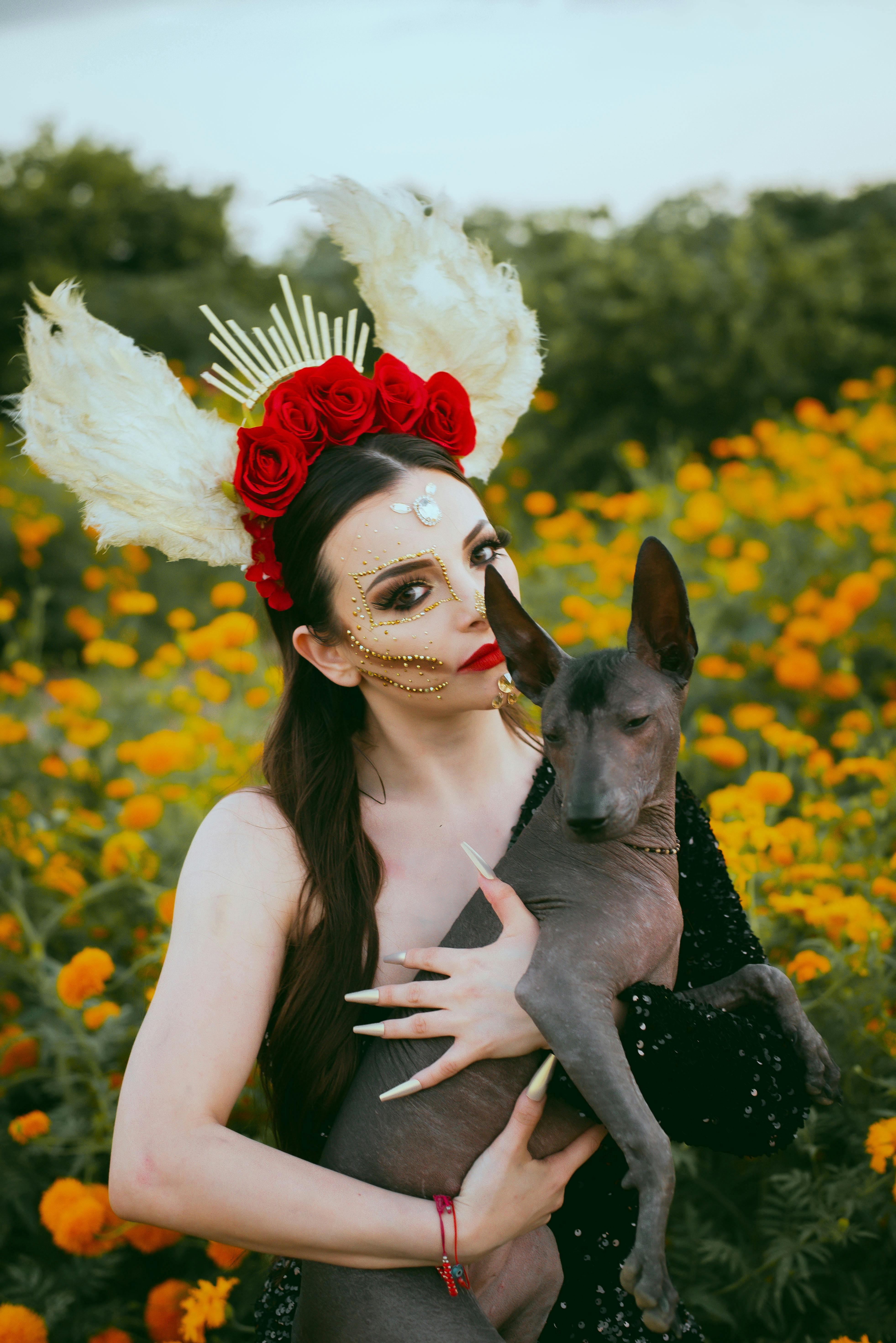 Woman in elaborate costume with flowers and feathers holds dog in a vibrant flower field.