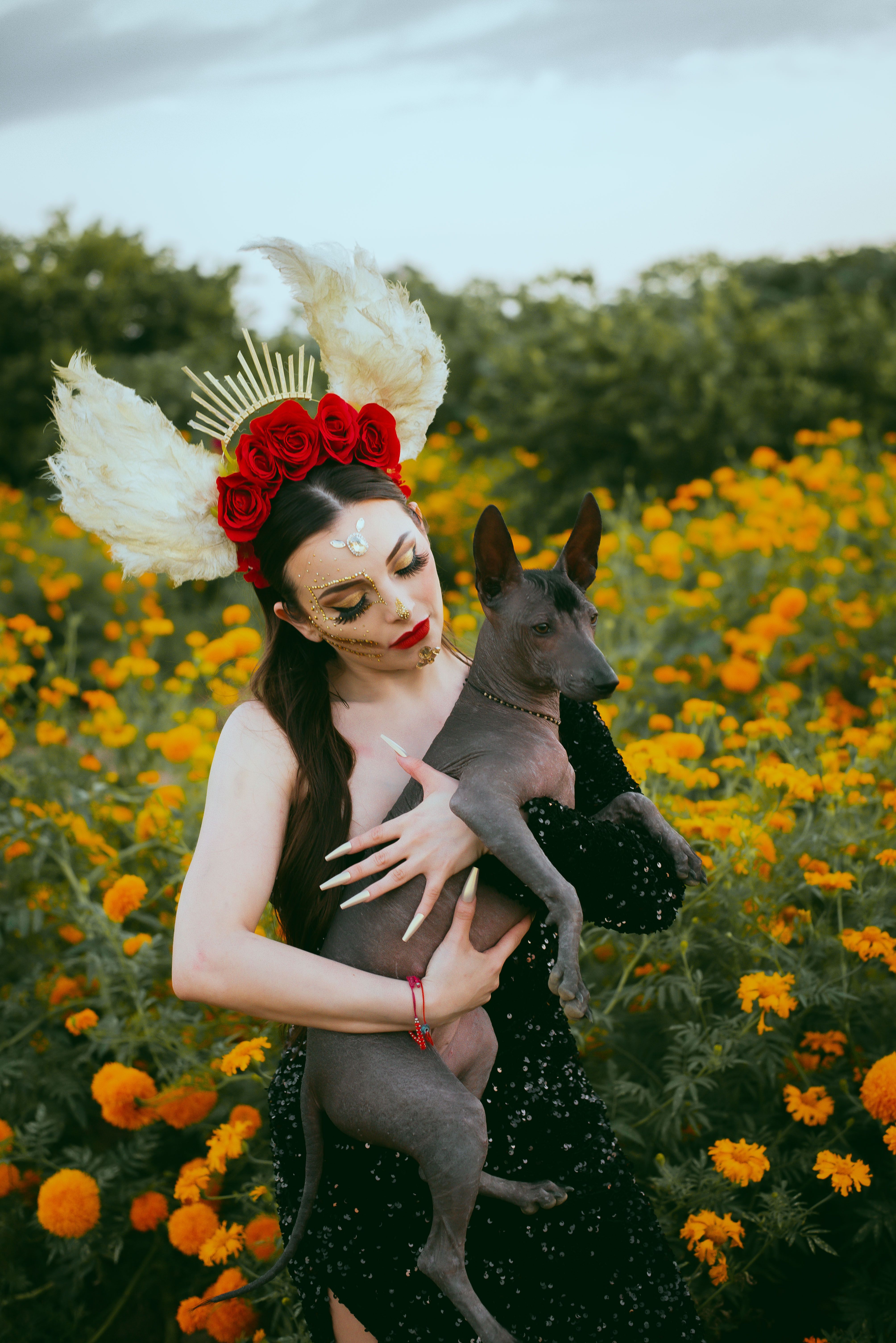 Artistic portrait of a woman in costume holding a Mexican hairless dog amidst vibrant marigolds.