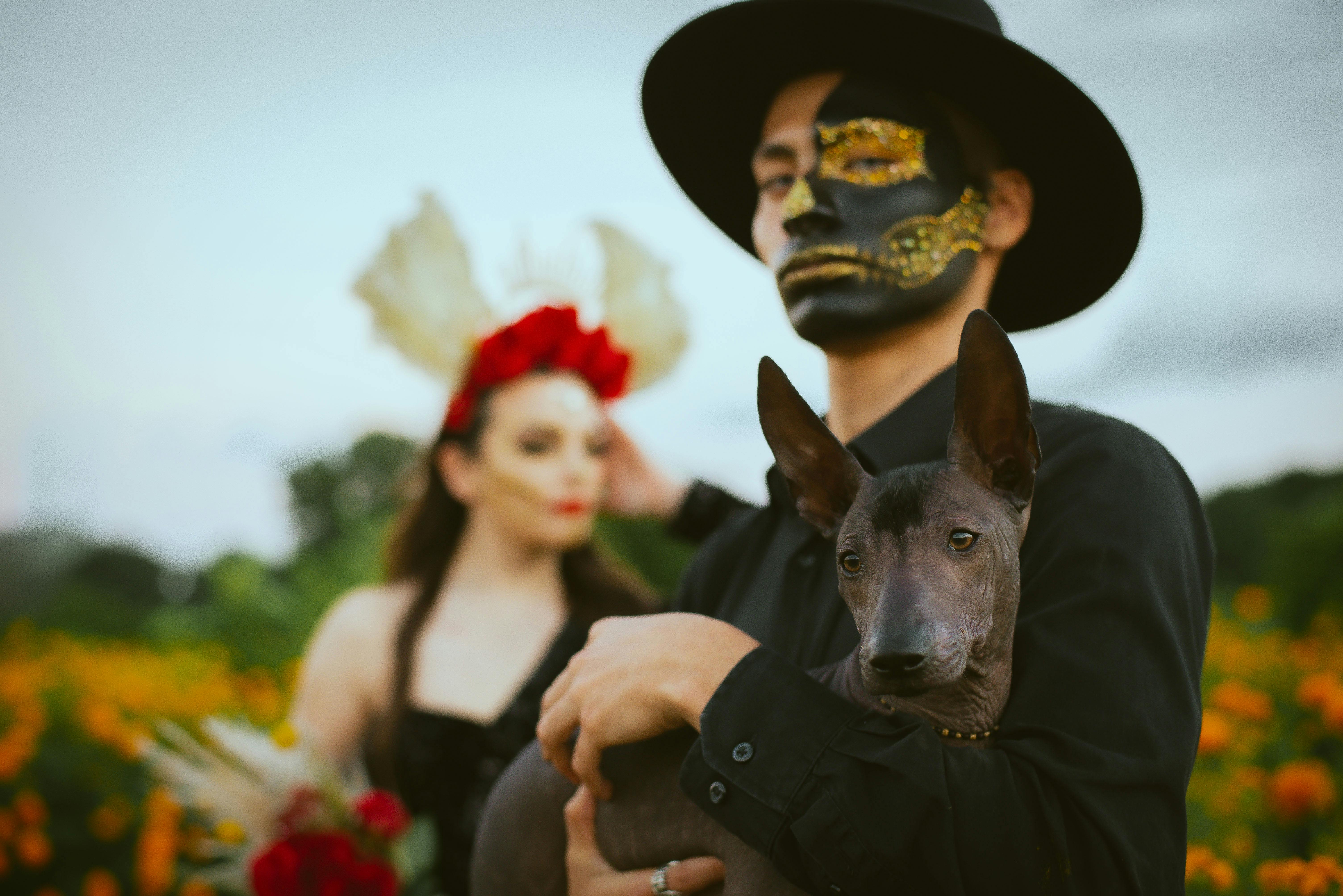 A man with artistic makeup holding a dog, embracing Day of the Dead themes in a floral setting.