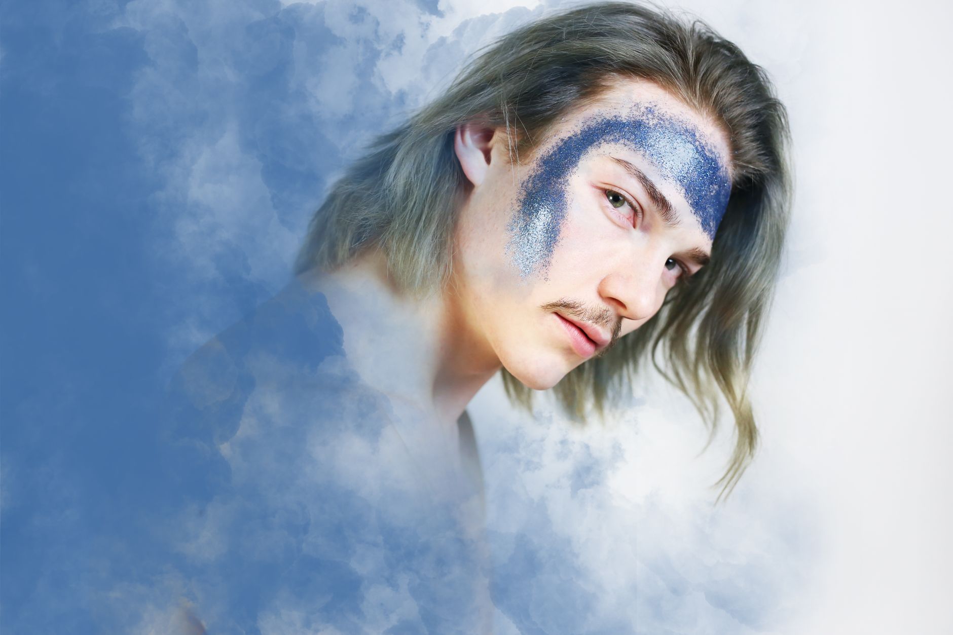 Artistic portrait of a young man with glitter face paint, set in a cloudy studio background.