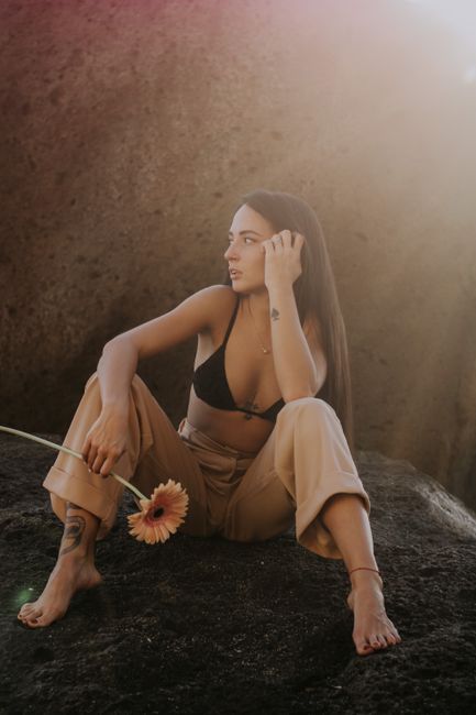 Woman in casual attire sitting on rocks holding a sunflower, captured in natural light.