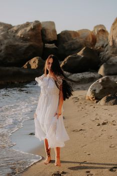 A woman in a flowing white dress strolls along a sandy beach near rock formations.