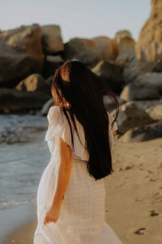 Woman in a flowing white dress walking on a rocky beach at sunset.