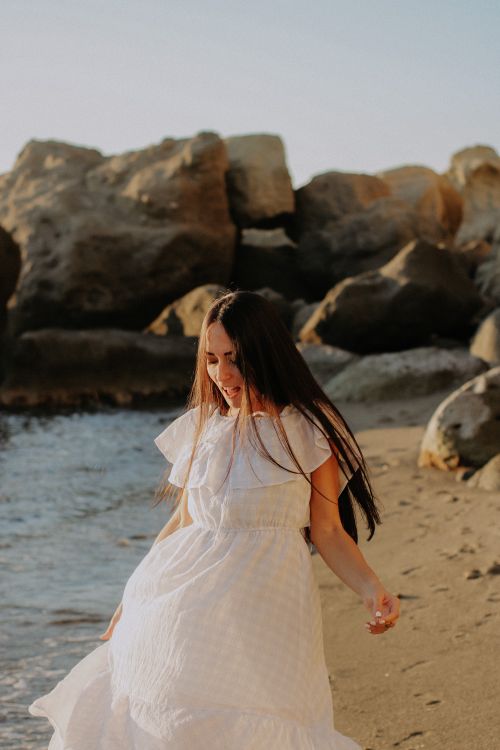 Free A woman in a white dress walks along a scenic beach with boulders. Stock Photo