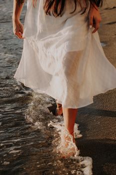A woman in a white dress walks barefoot along the beach, touching the waves.