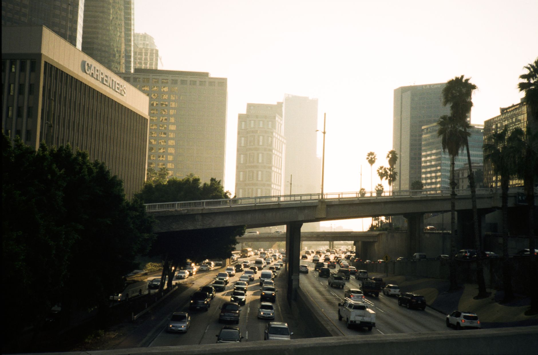 Heavy traffic on a Los Angeles freeway at sunset, with the downtown skyline and palm trees behind