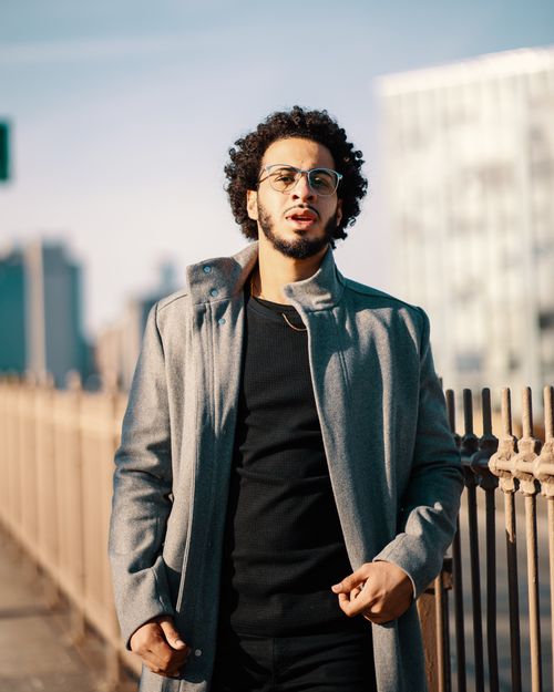 Free An African American man in glasses and a gray coat poses stylishly on an urban street. Stock Photo