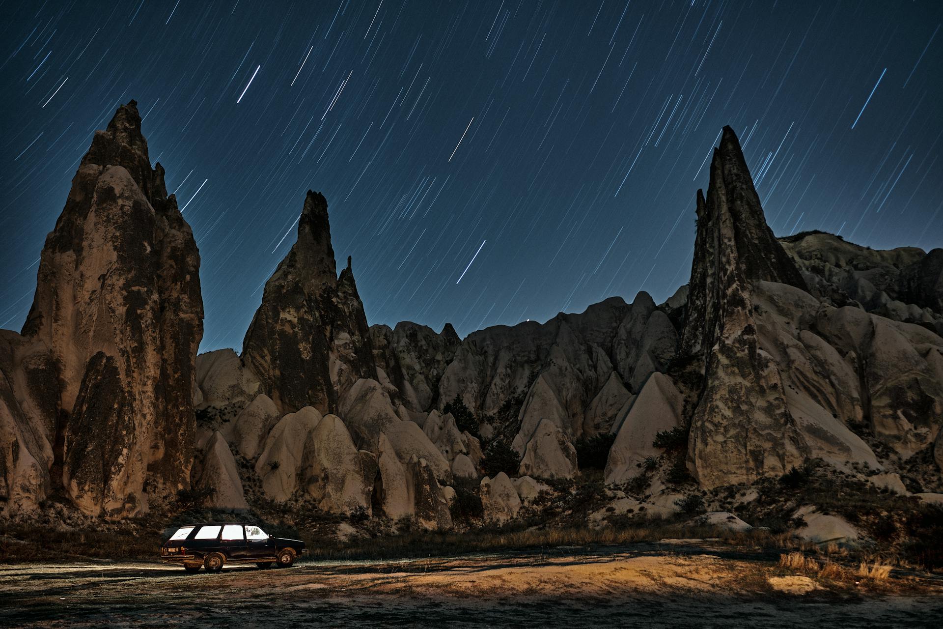 Long exposure of star trails over dramatic rock formations and a car under a starry sky.