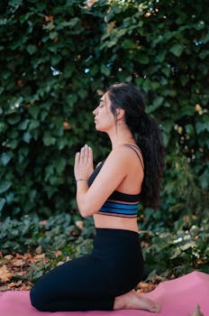 Side view of a woman meditating on a yoga mat outdoors, surrounded by lush greenery.