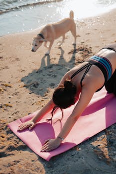 Woman performing yoga on a beach mat with a dog nearby on a sunny day.