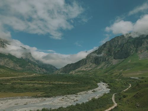 Free Stunning aerial view of a lush green valley bordered by rugged mountain ranges and a winding river under a blue sky. Stock Photo