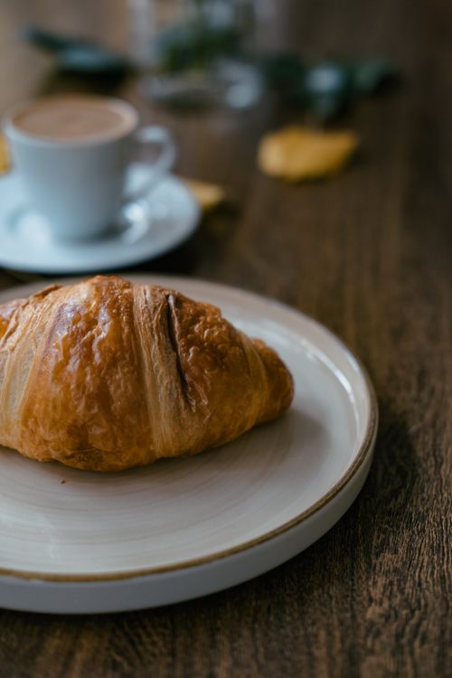 Free A beautifully baked croissant on a ceramic plate with a blurred coffee cup in the background. Stock Photo