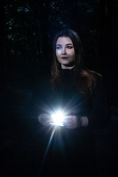 Woman in a dark forest holding a bright light, creating a magical aura.