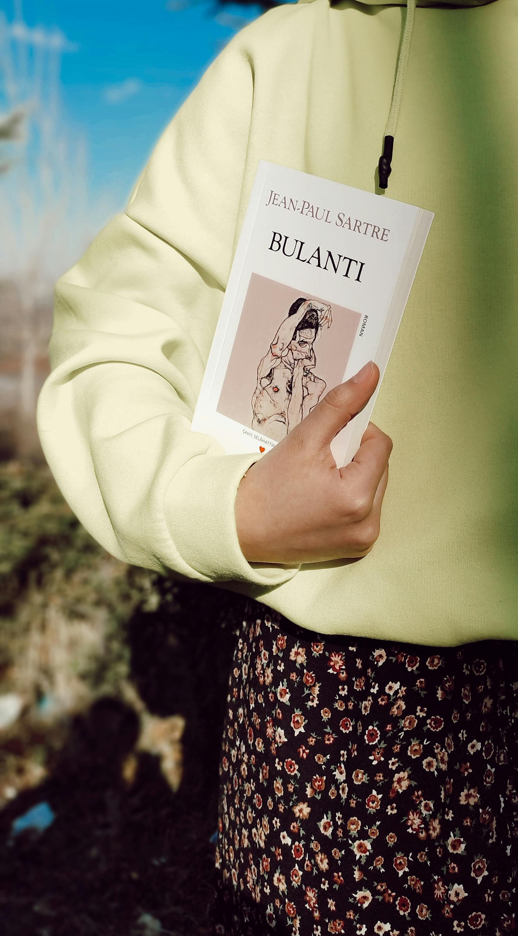 Close-up of person holding 'Bulantı' by Jean-Paul Sartre outdoors, wearing a yellow sweater.
