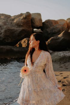 A woman in a floral dress stands by a rocky beach, holding a flower, near the shore.