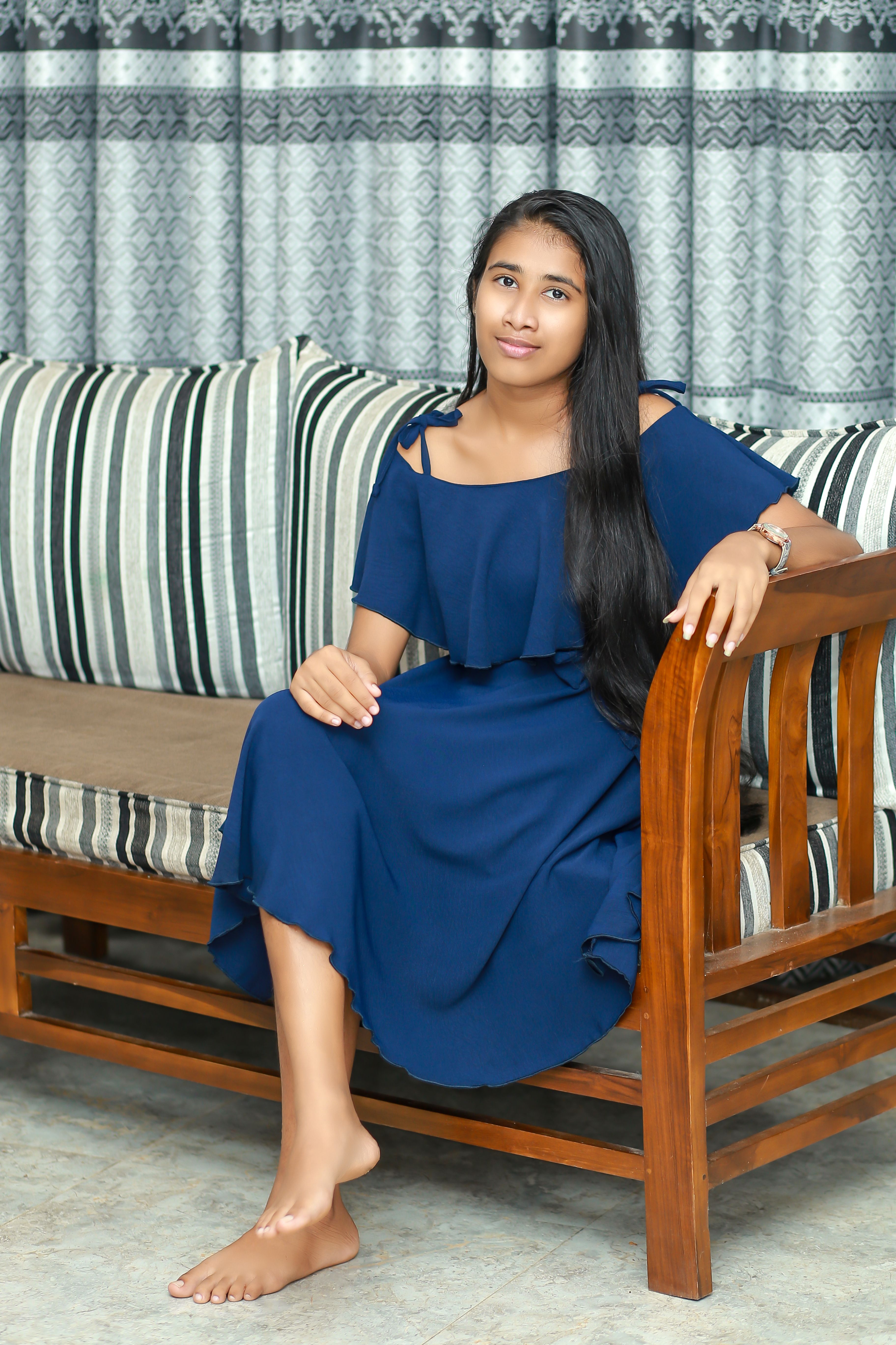 Woman wearing a blue dress sits elegantly barefoot on a wooden sofa indoors.