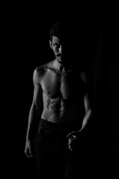 Grayscale image of a shirtless man standing against a black background, exuding a dramatic and moody atmosphere.