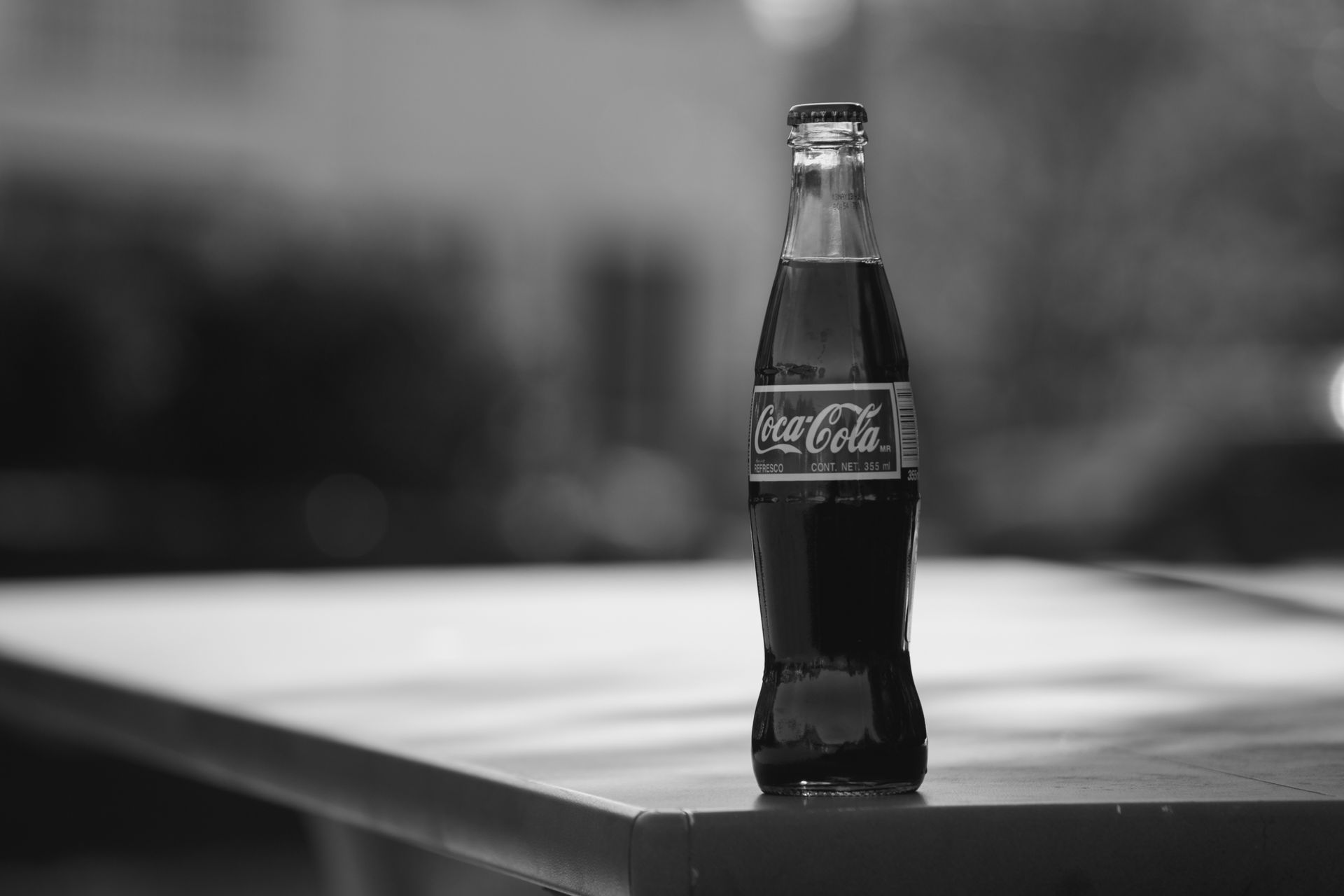 Black and white close-up of a classic Coca-Cola bottle on a table outdoors.
