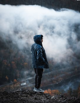 Person in a blue jacket standing on a misty cliff, embracing nature. Captured in the wild outdoors.