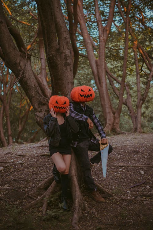 Two People Standing Beside a Tree Wearing Carved Pumpkins on Head 