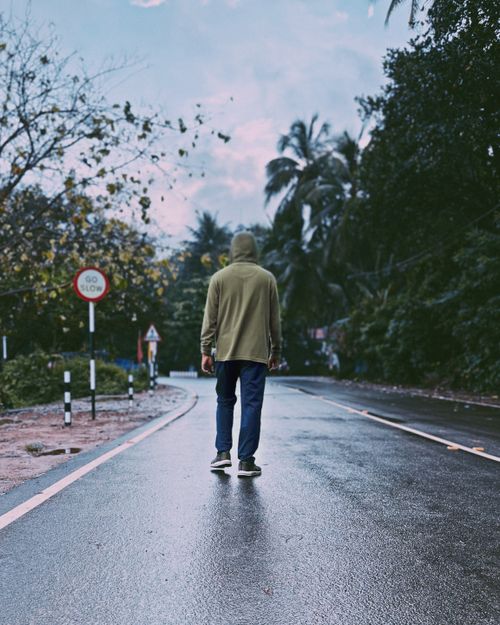 Free A person in a hoodie walking alone down a wet road in Kochi, India, surrounded by lush foliage. Stock Photo