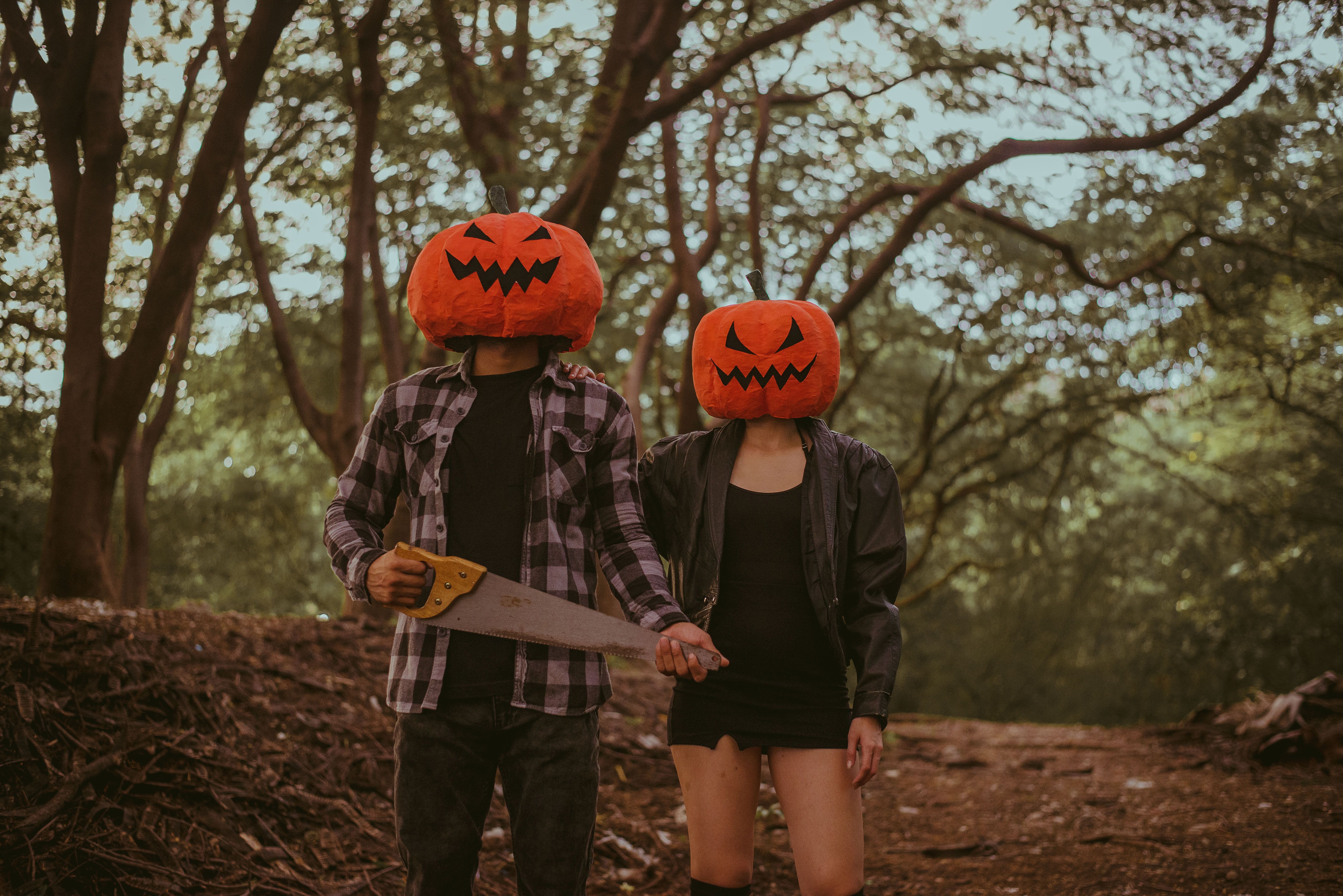 Two people wearing pumpkin head costumes and holding a saw in a spooky forest setting.