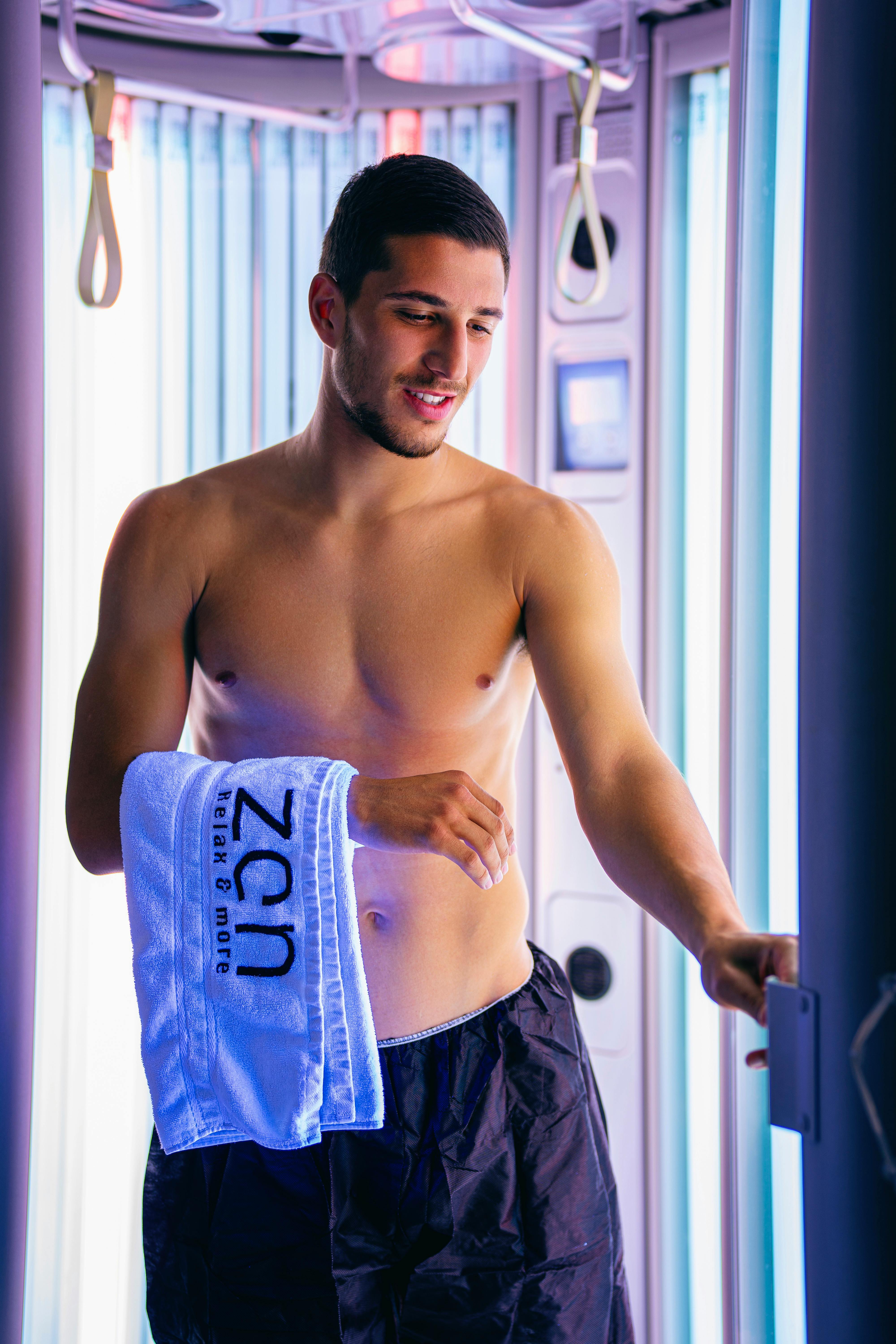 Shirtless young man with towel smiling in a modern tanning booth.