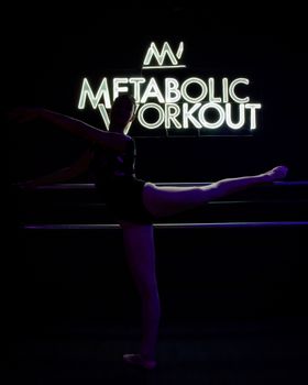 Silhouette of a ballet dancer stretching in a neon-lit gym with 'Metabolic Workout' sign.