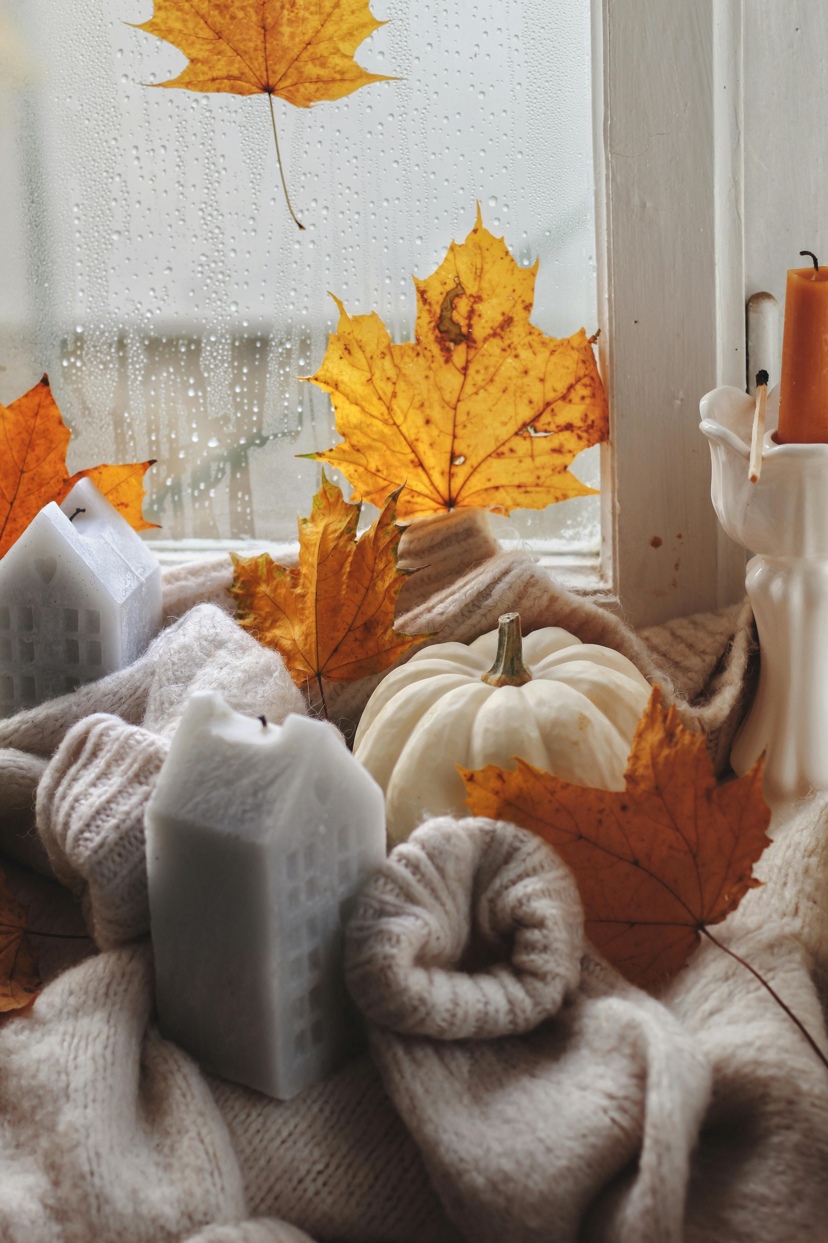 Warm autumn setup with leaves, pumpkin, and candles on a rainy windowsill.
