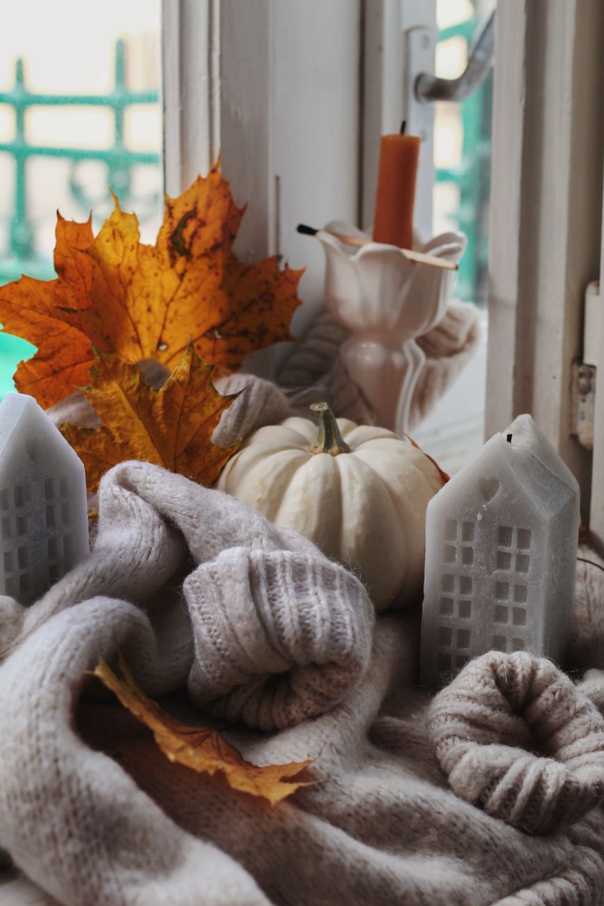Warm and cozy autumn setting featuring a white pumpkin, candles, and fall leaves by a window.