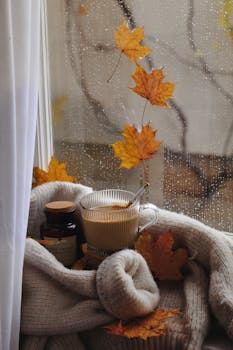 Warm coffee next to autumn leaves on a rainy windowsill evokes cozy fall vibes.