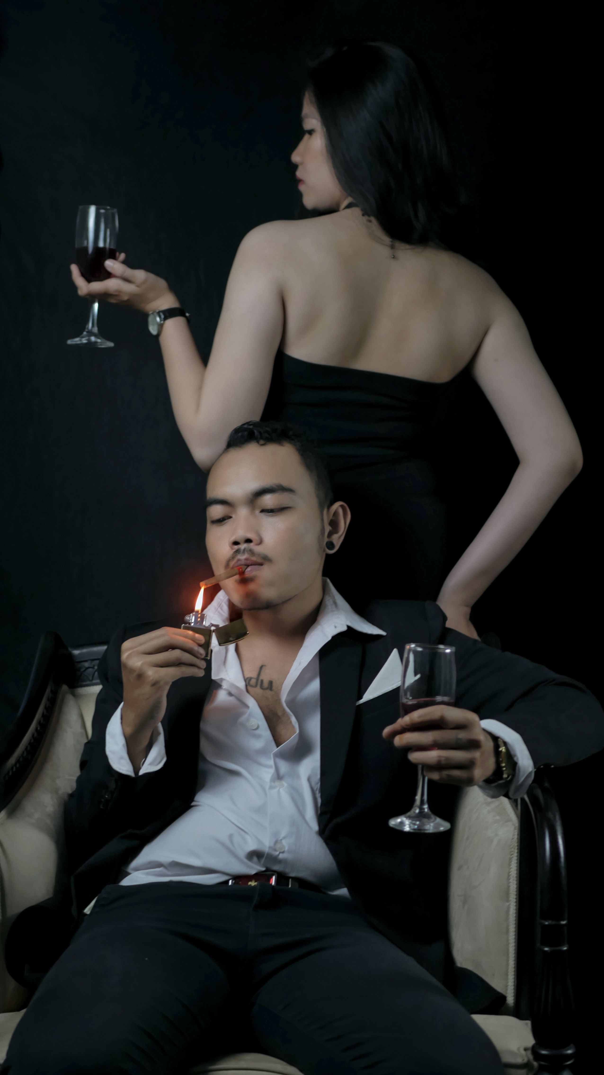 A fashionable couple in sophisticated attire enjoying wine and cigars indoors, exuding style and elegance.